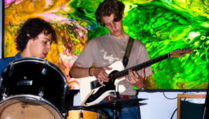 Een jongere achter de drums en een jongere op gitaar muziek aan het maken tijdens de jamsessie