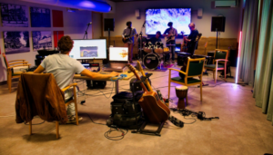 Overzichtsfoto van de jamsessie. We kijken de producer op de rug, verderop op het podium staan drie gitaristen en een drummer